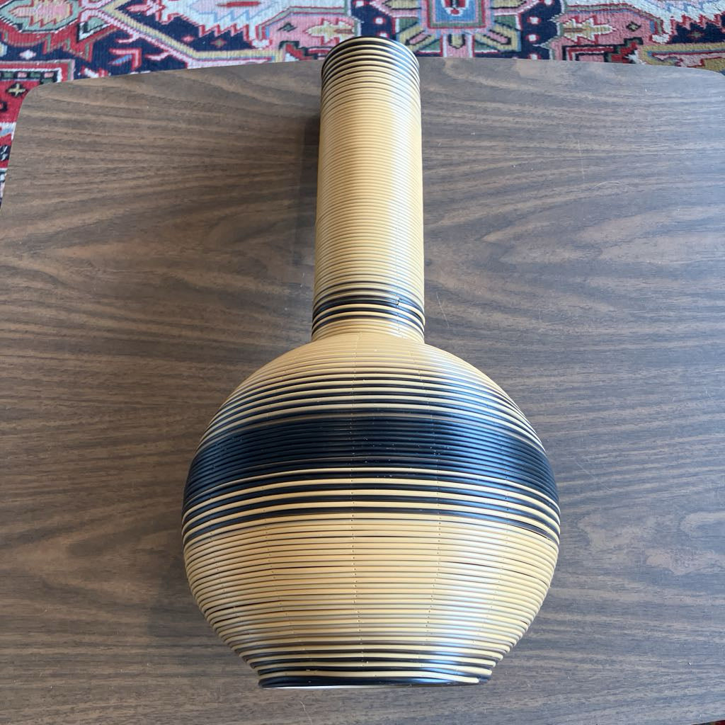 A hand-woven decorative vase with a bulbous body and a long neck, made from natural bamboo or wood strips with black painted accents, displayed on a dark wood tabletop.