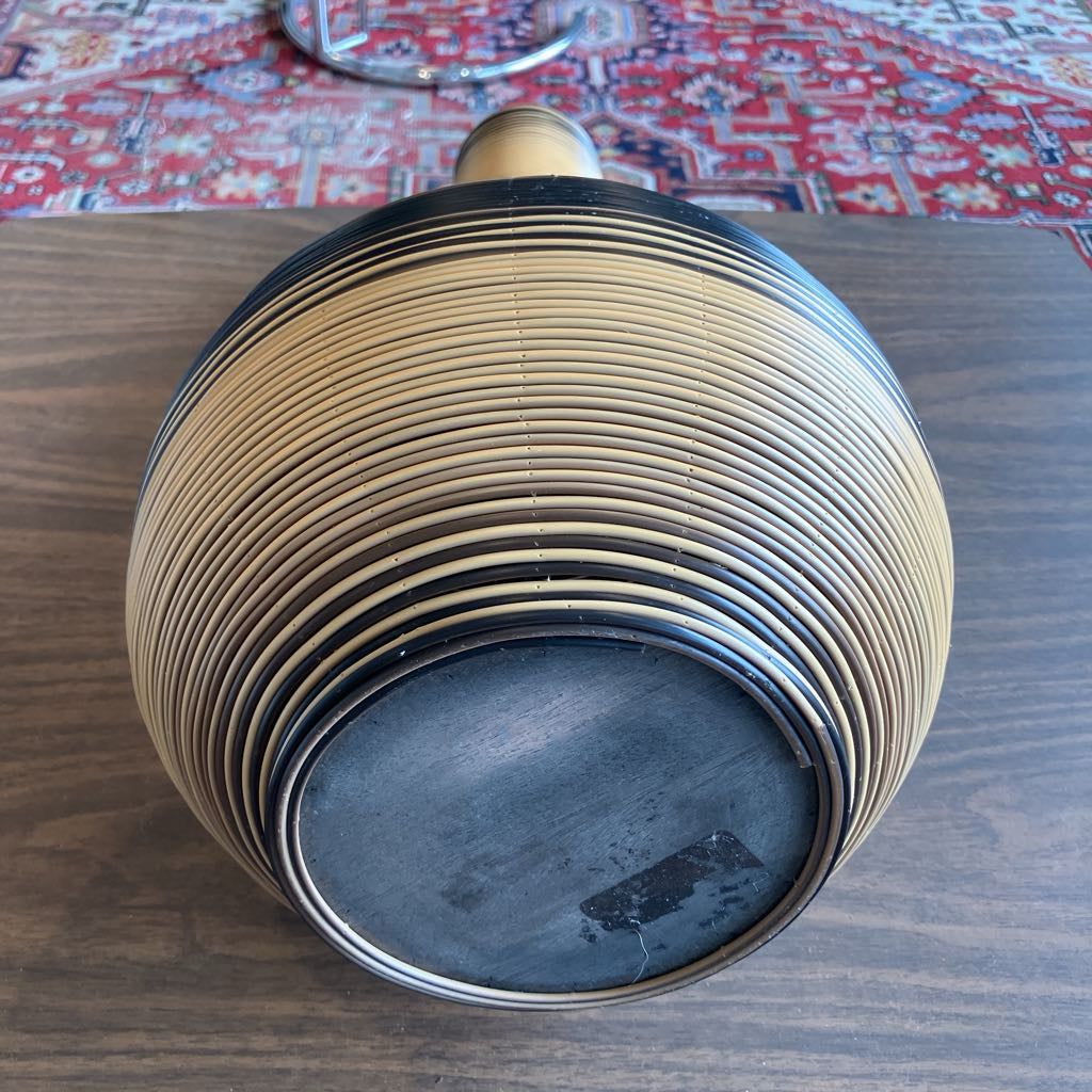 A round wooden decorative bowl or vase with a ribbed, striped pattern in natural tan and black, photographed on a dark wood tabletop with a patterned red rug in the background.