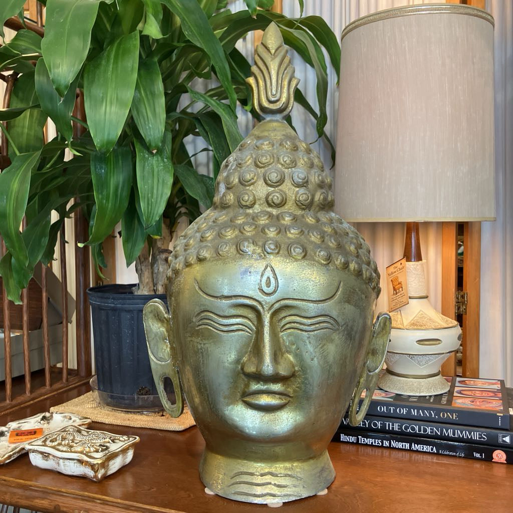 A large decorative Buddha head sculpture with a distressed metallic gold finish, displayed on a wooden table in a home setting next to a potted plant, lamp, and books.