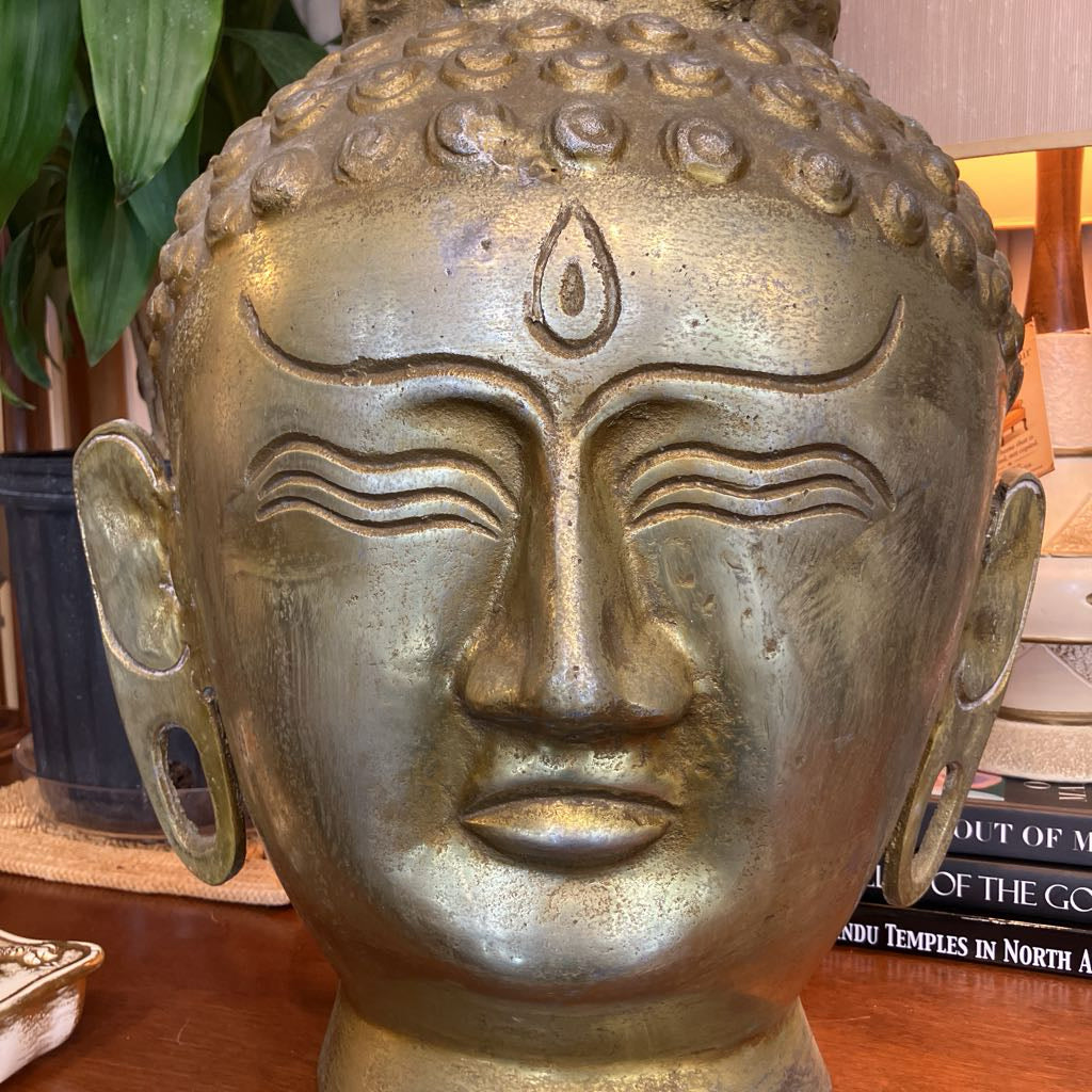 A large metallic Buddha head sculpture with an antique bronze finish, displayed on a wooden surface with books and a plant in the background.