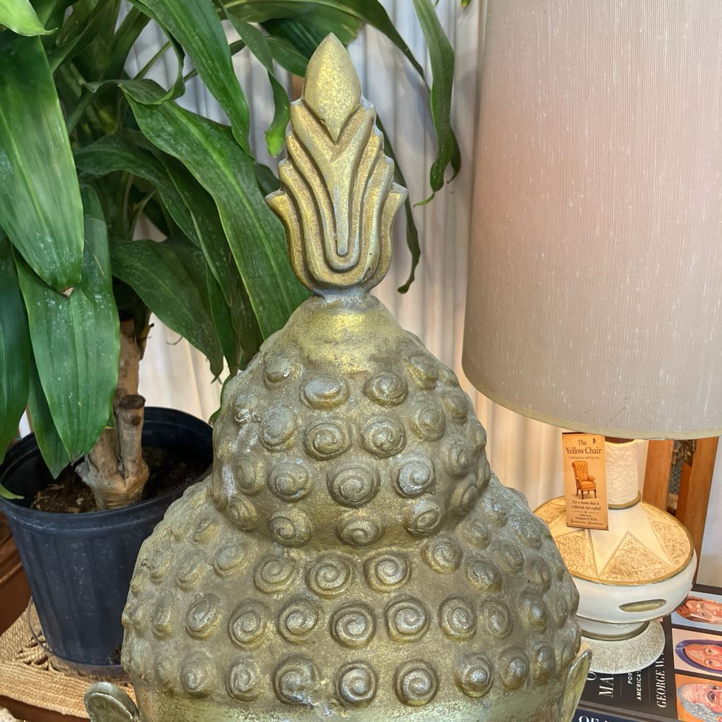 The top of a buddha statue with the head resembling a stylized lotus or lotus bud, finished in a distressed metallic gold/bronze color. It's styled in a home environment next to a potted green plant and a beige fabric lampshade.