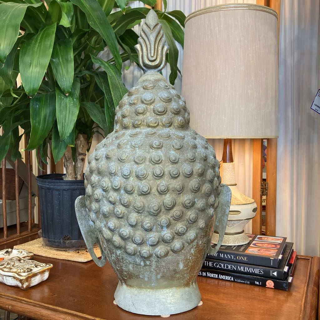 A large decorative Buddha head sculpture with a textured, bubble-covered surface in a muted sage green-gray finish, displayed on a wooden table in a home setting next to a potted plant, a table lamp, and books.