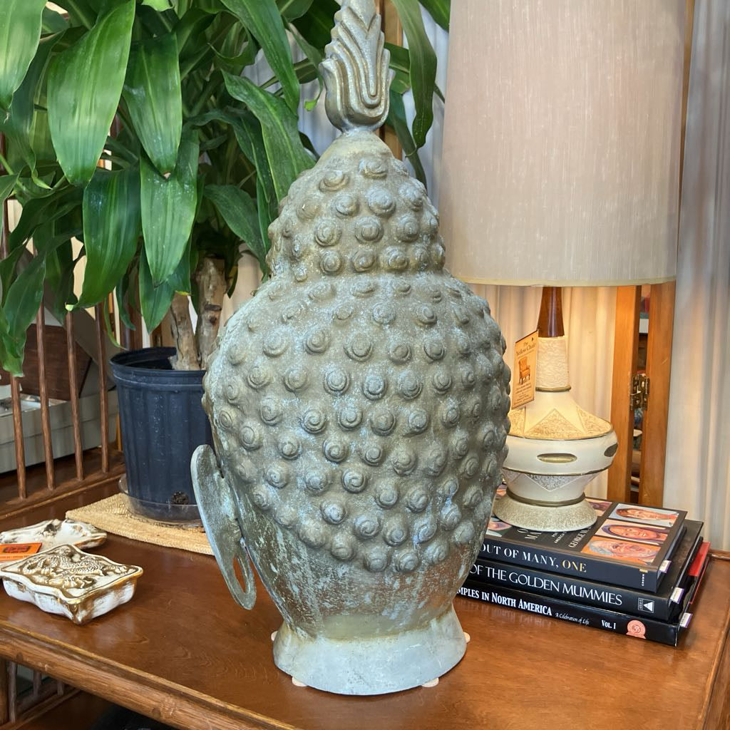 A large decorative Buddha head sculpture with a textured, bumpy surface and a pointed top, displayed on a wooden table in a home setting next to a potted plant, books, and a lamp.