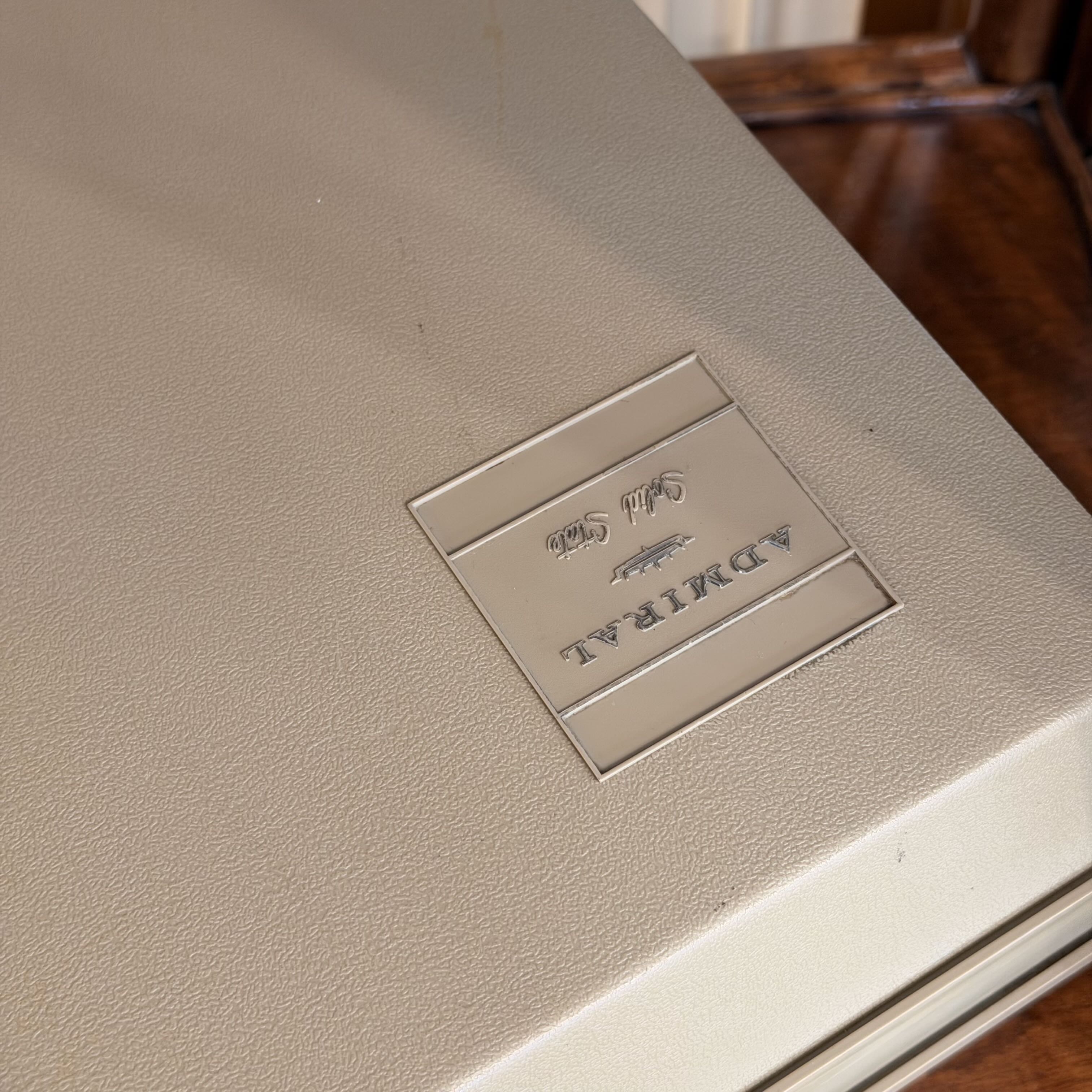 Close-up of a beige textured hard case with a silver metal plaque embossed with the brand name ADMIRAL.