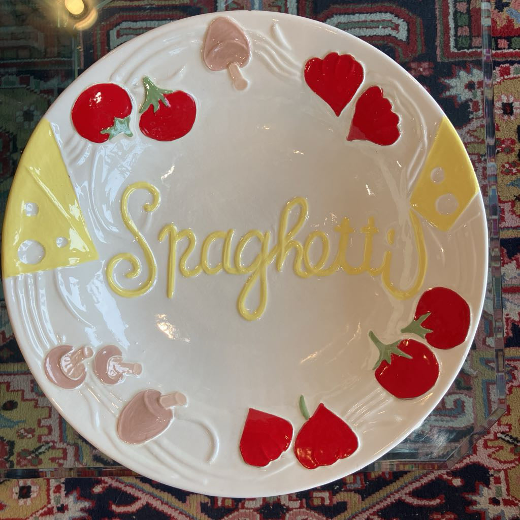 A white ceramic decorative plate featuring the word 'Spaghetti' in yellow script, surrounded by hand-painted red tomatoes, yellow cheese wedges, and pink mushrooms, set on a patterned rug.