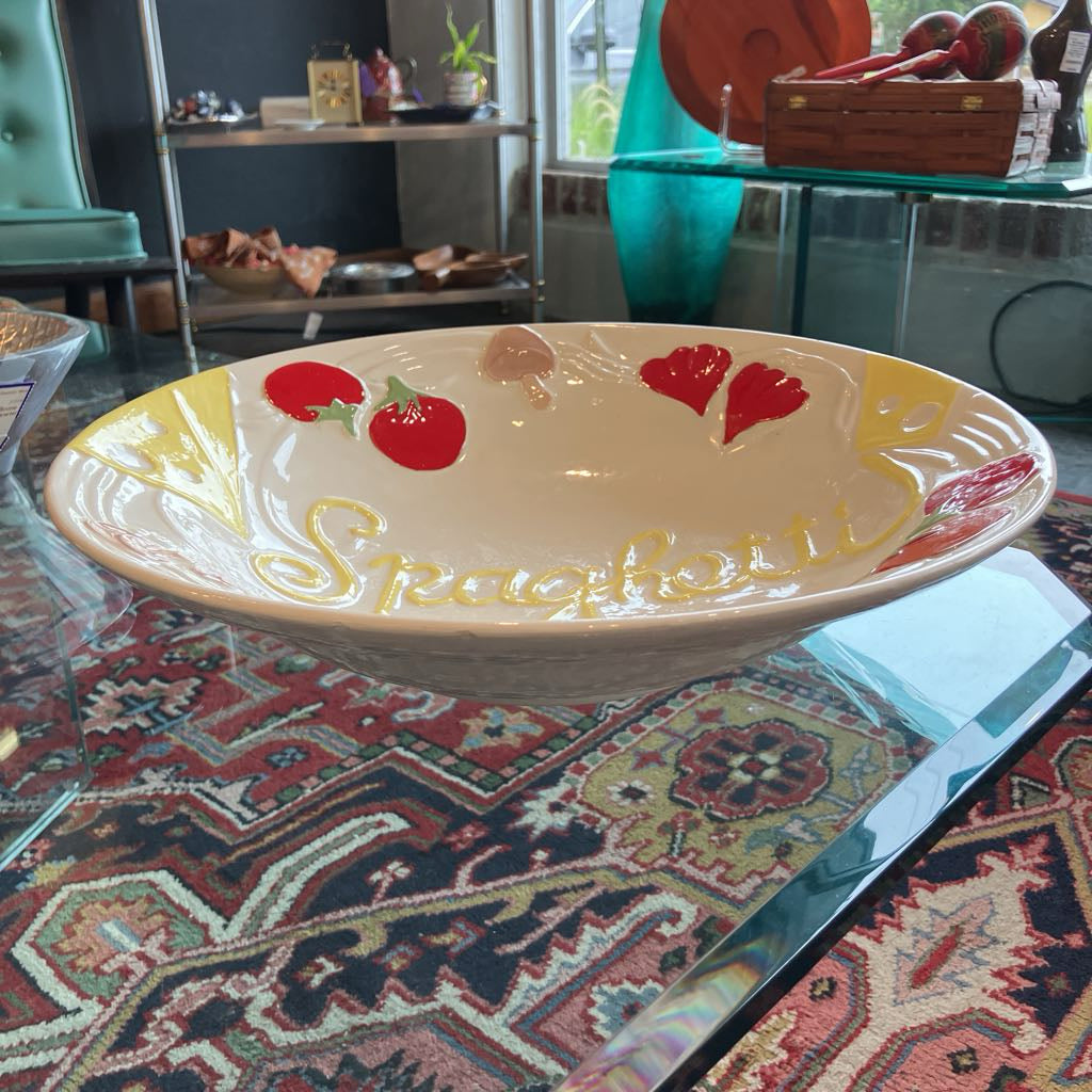 A wide, shallow ceramic bowl with a cream-colored interior, decorated with red strawberry motifs, yellow 'Spaghetti' script, and red heart details, resting on a patterned rug in a home setting.