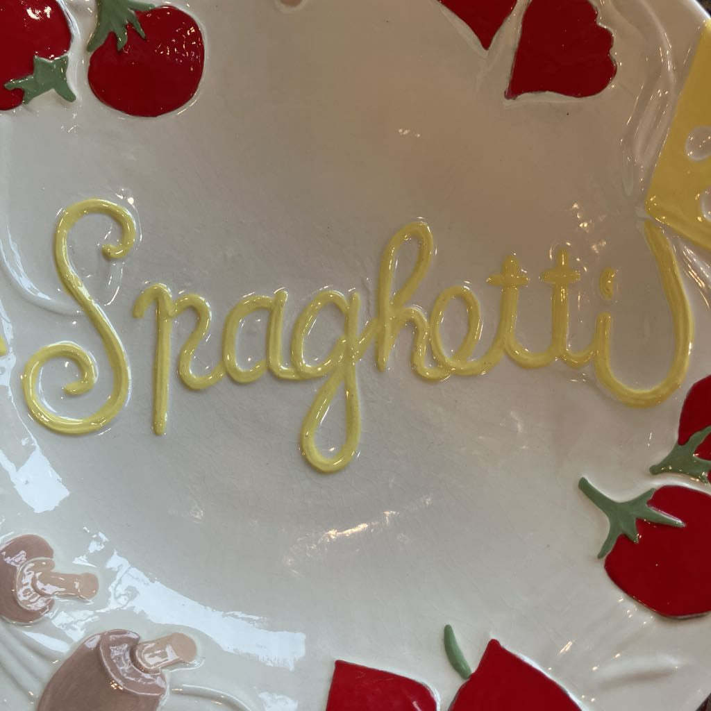 A close-up of a white ceramic plate featuring the word 'Spaghetti' written in yellow icing-like lettering, surrounded by red and green strawberry motifs.