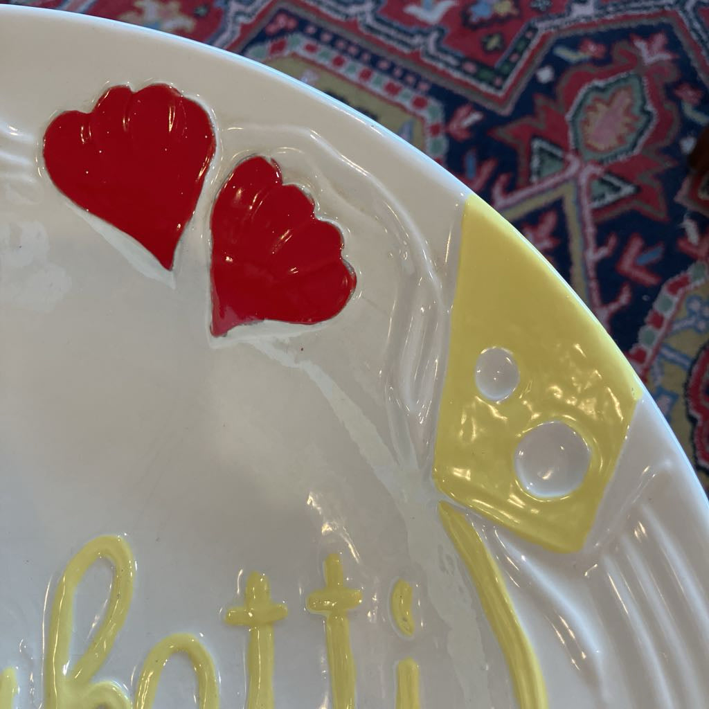 Close-up of a white ceramic plate with two red hearts, a yellow cheese wedge with white dots, and yellow script lettering on the rim, set against a patterned rug.