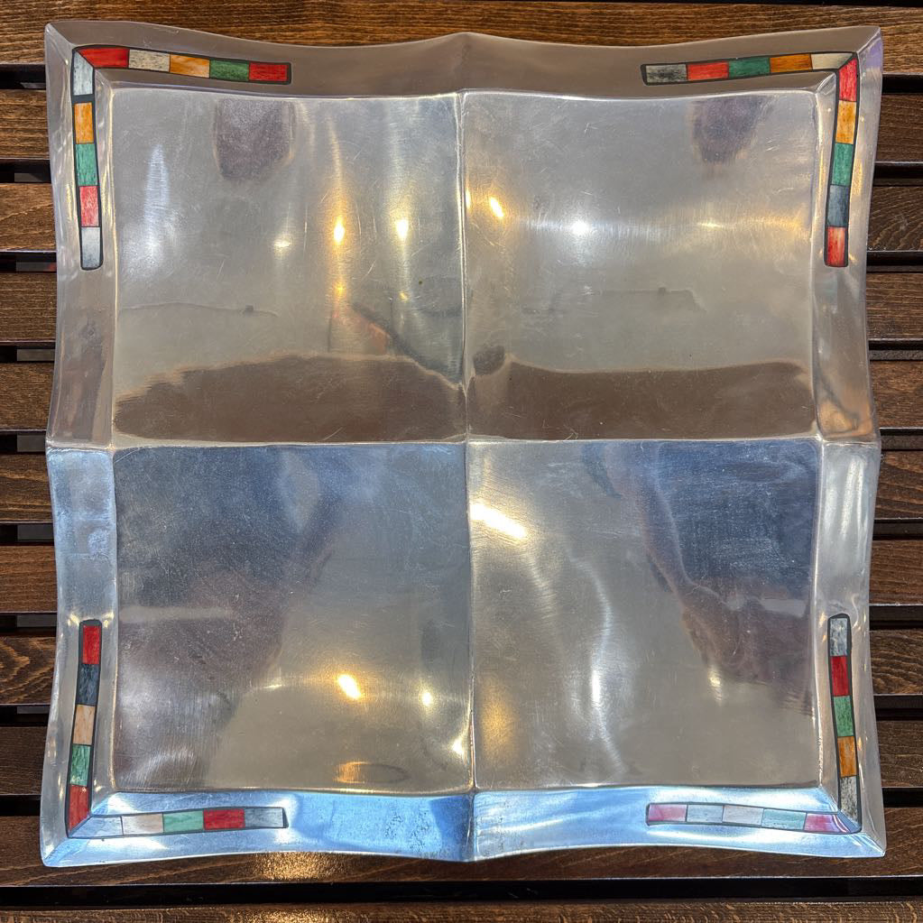 A large aluminum tray with square curved segments and mosaic inlaid corners on a wooden surface.