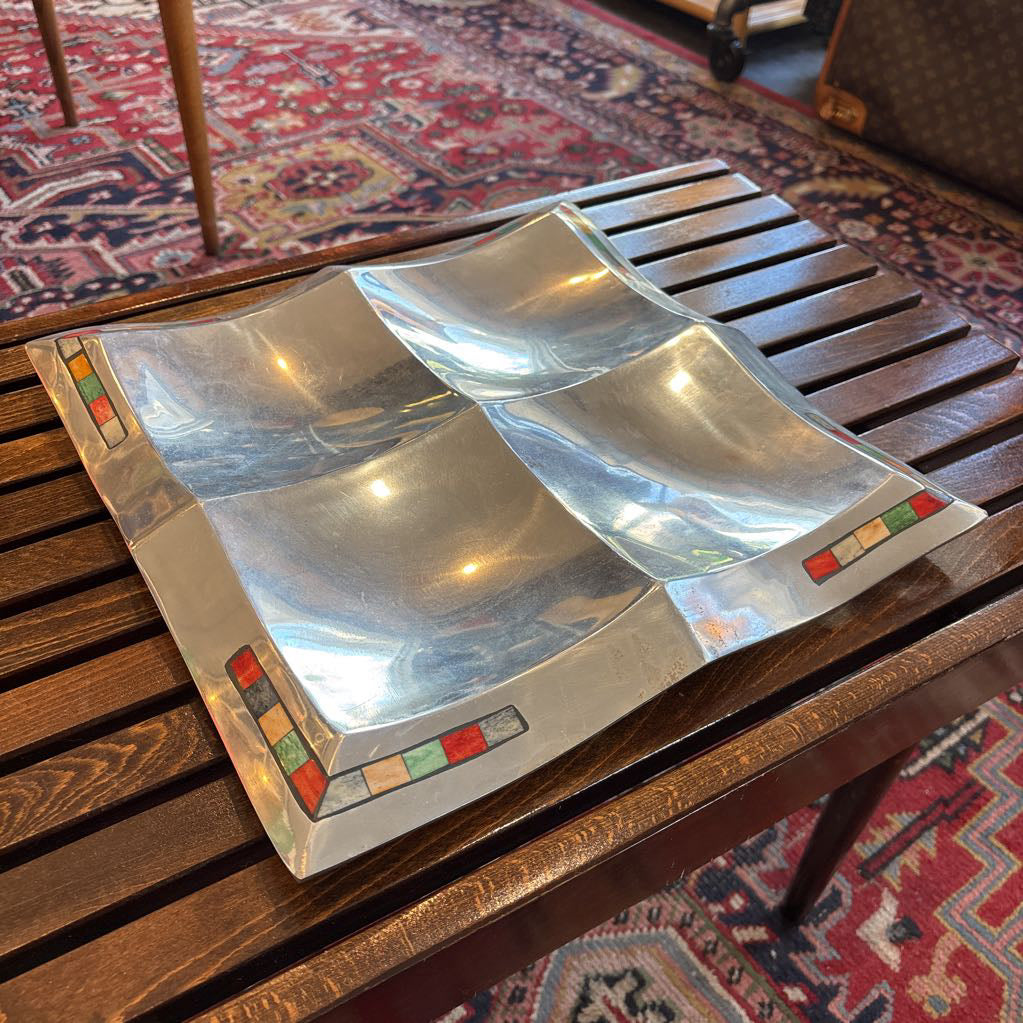 A large aluminum tray with square curved segments and mosaic inlaid corners on a wooden surface.