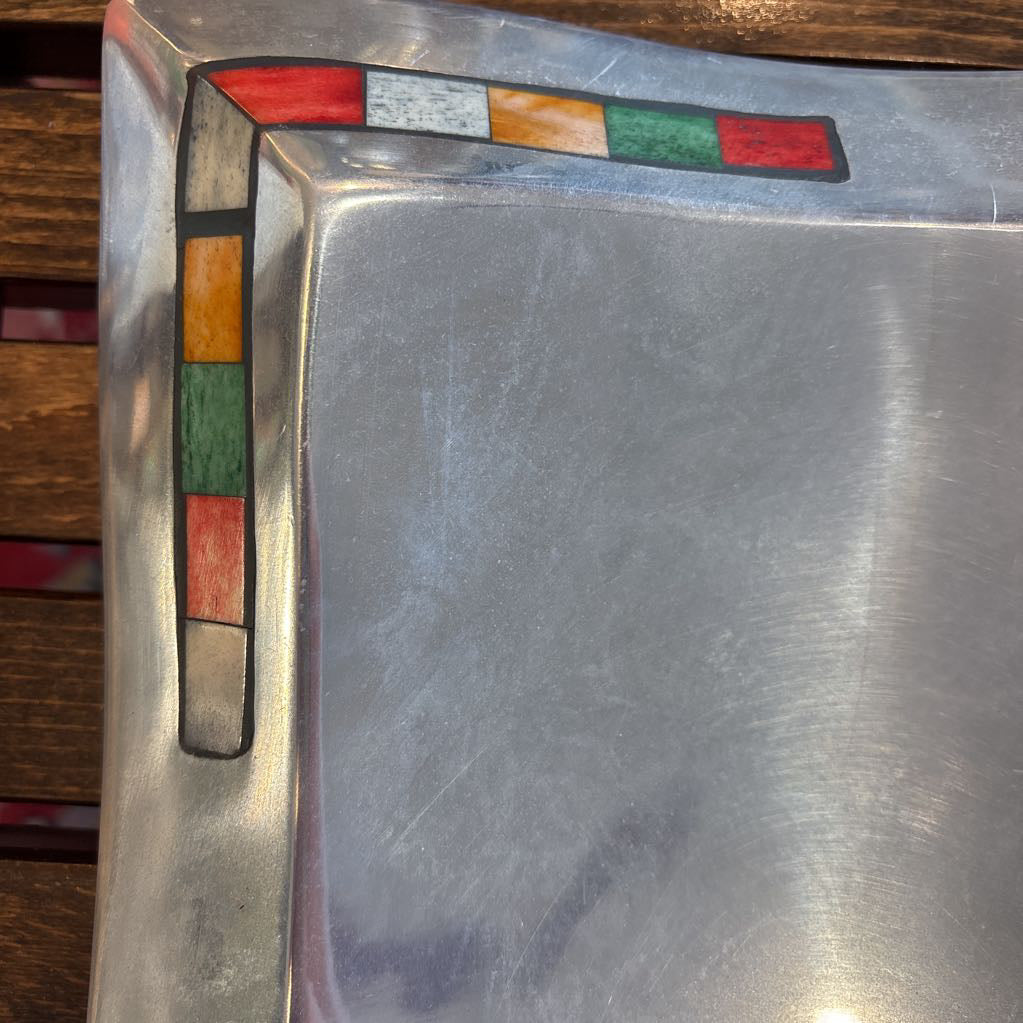 A large aluminum tray with square curved segments and mosaic inlaid corners on a wooden surface.