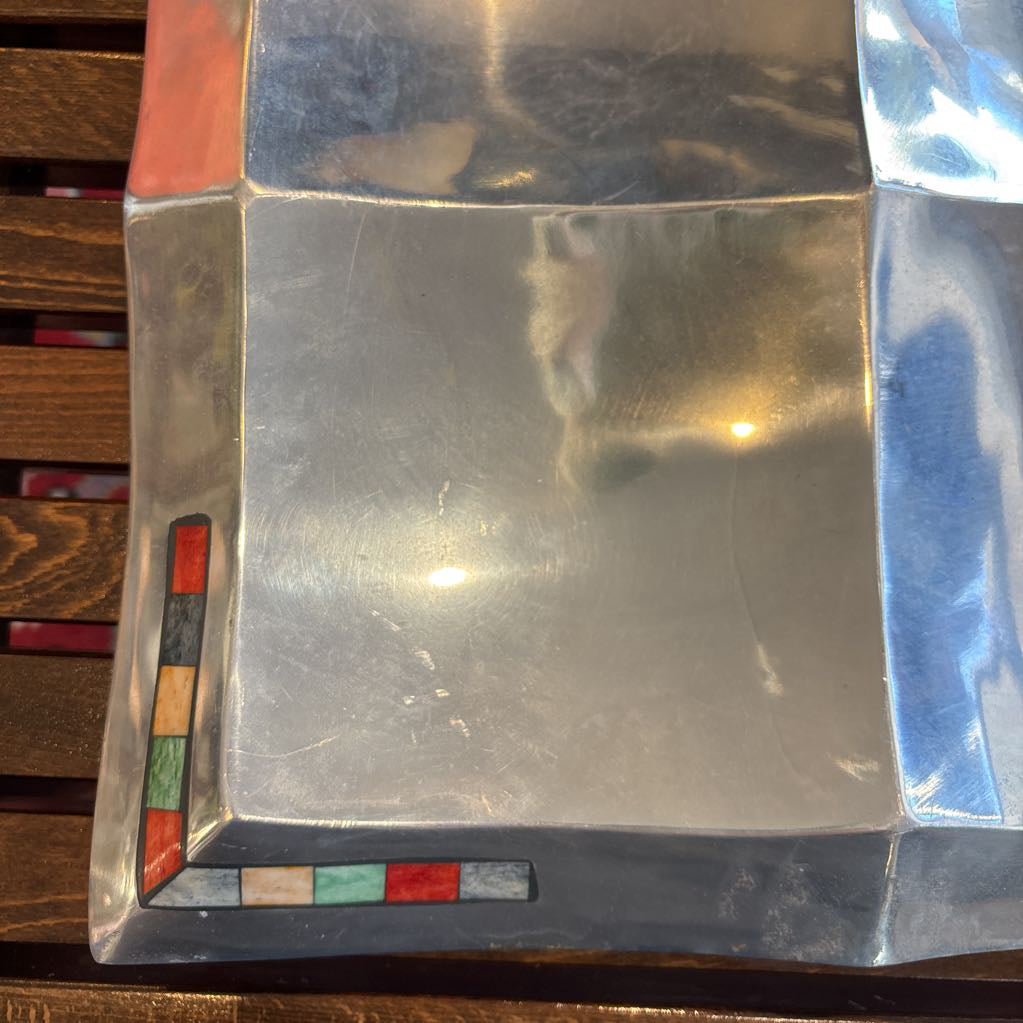 A large aluminum tray with square curved segments and mosaic inlaid corners on a wooden surface.