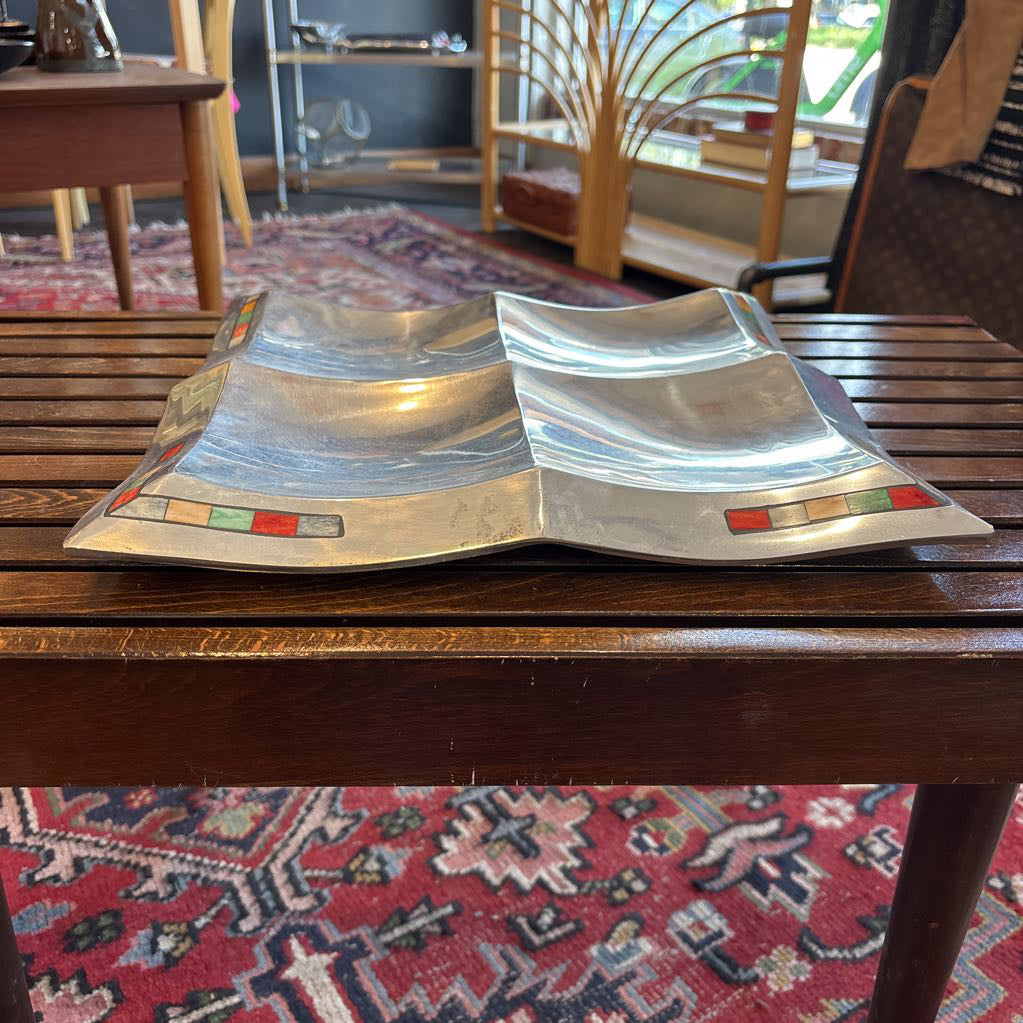 A large aluminum tray with square curved segments and mosaic inlaid corners on a wooden surface.