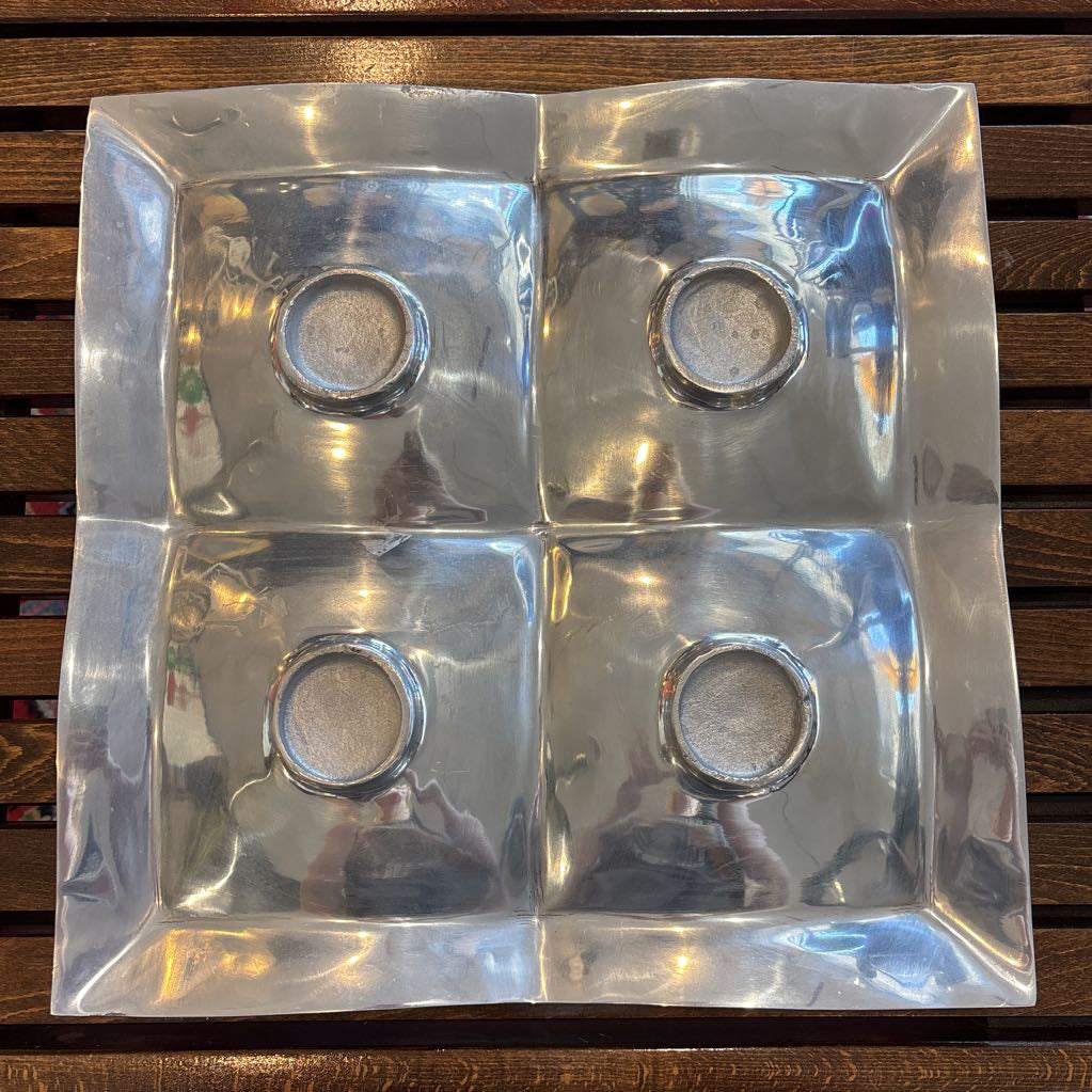 A large aluminum tray with square curved segments and mosaic inlaid corners on a wooden surface.