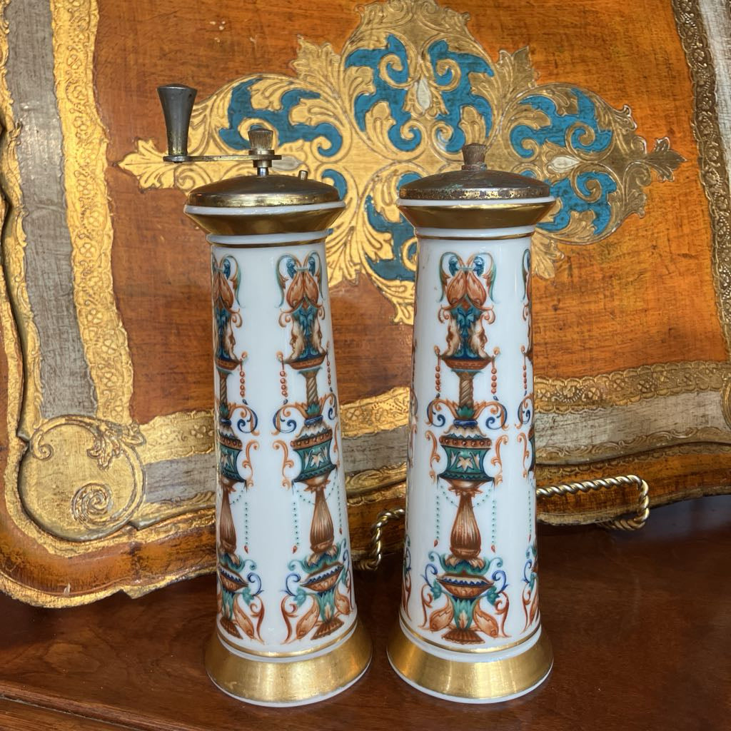 A pair of vintage ceramic pepper mills with gold-toned metal caps and handles, featuring a white porcelain body decorated with a blue and brown urn pattern, set against an ornate gold and blue tapestry background.