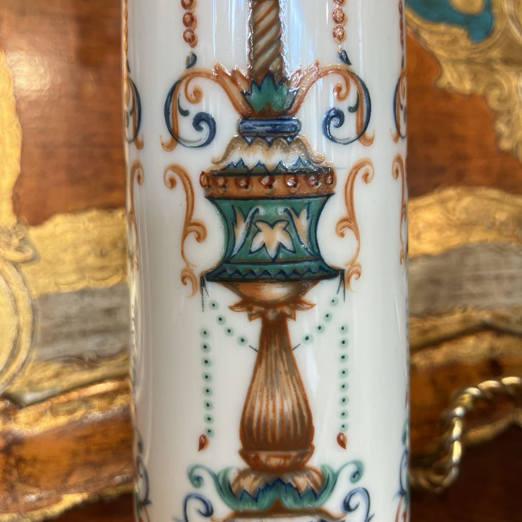 Close-up of a white porcelain vase featuring a hand-painted classical urn motif in teal, gold, and brown, set against a background of ornate gold and brown wood paneling.