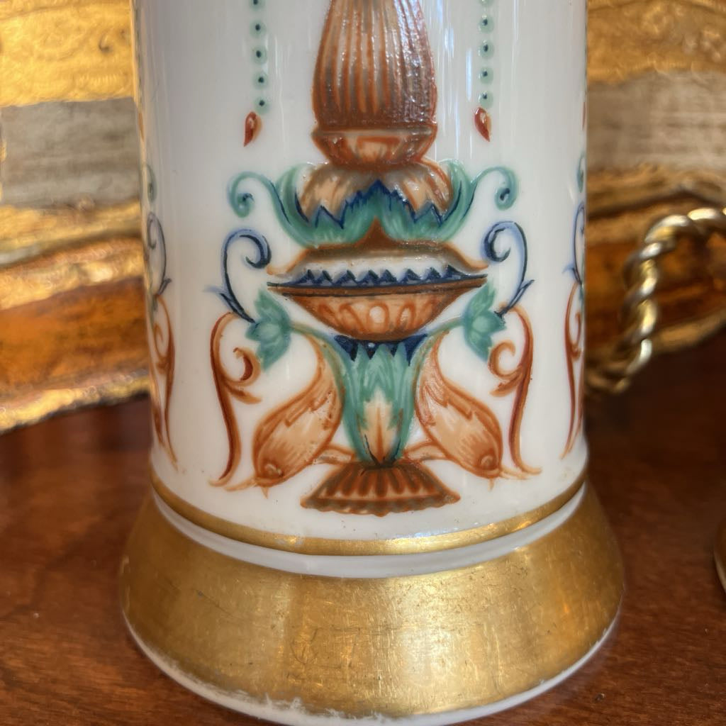 A close-up of a white ceramic mug featuring a hand-painted brown and turquoise urn motif with gold-gilded detailing on the base, set on a wooden surface.