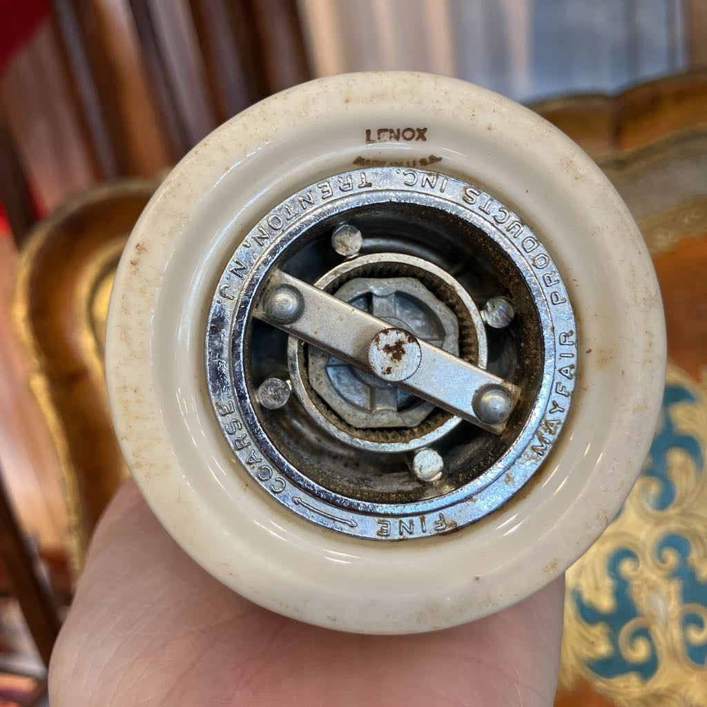 A close-up of a vintage Lenox pepper mill, held in a hand. The circular cream-colored ceramic body shows the brand name 'LENOX' at the top, and the metal infuser assembly with brass pins and a rusted center nut is visible.