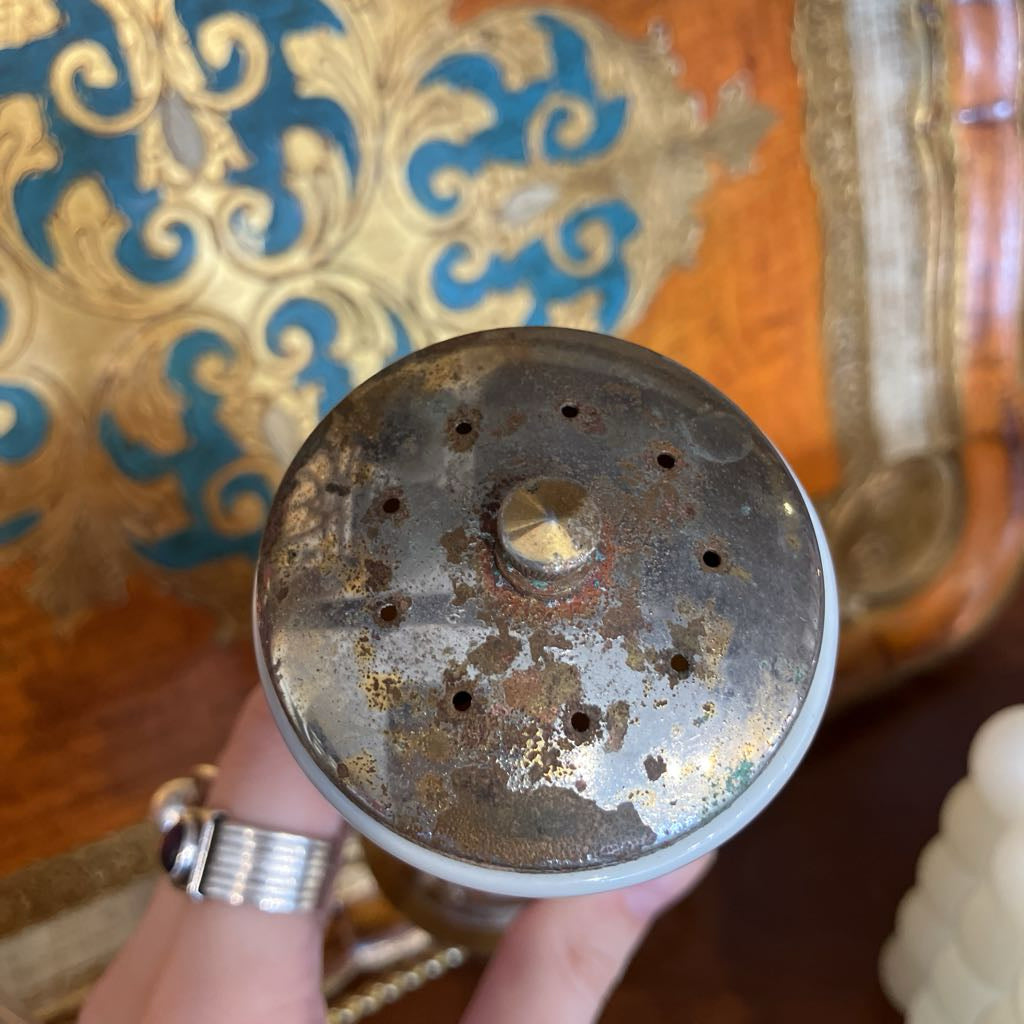 A close-up of a vintage metal spice or pepper grinder top, held by a hand. The top features a heavily weathered, rusted silver-toned metal surface with multiple small perforations and a central knob.