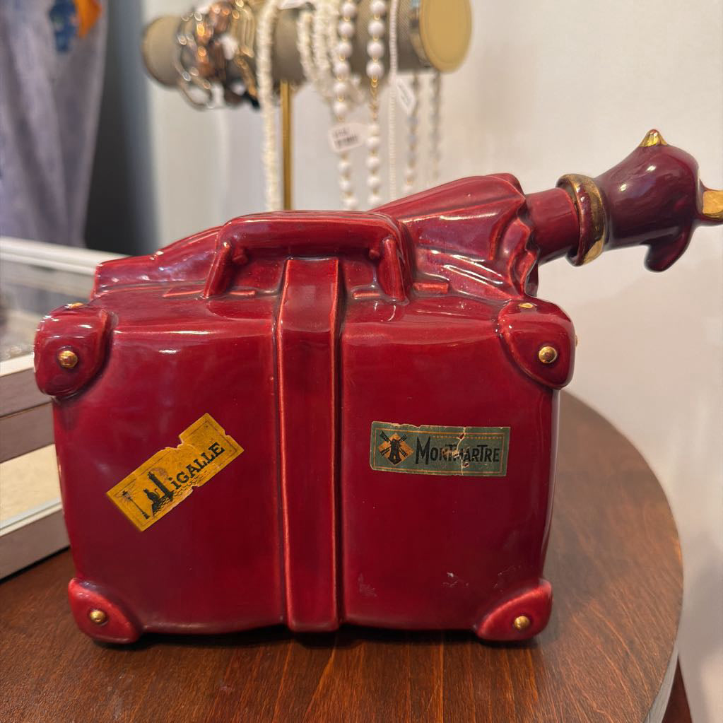 A glossy red ceramic or porcelain miniature suitcase-shaped object with gold-tone hardware, a yellow luggage tag reading "JACQUELINE," and a small plaque on the front, displayed on a wooden surface with jewelry in the background.