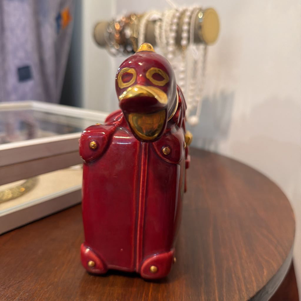 A glossy red duck figurine styled as a military officer in a mask, standing on a dark wooden table with a blurred background.