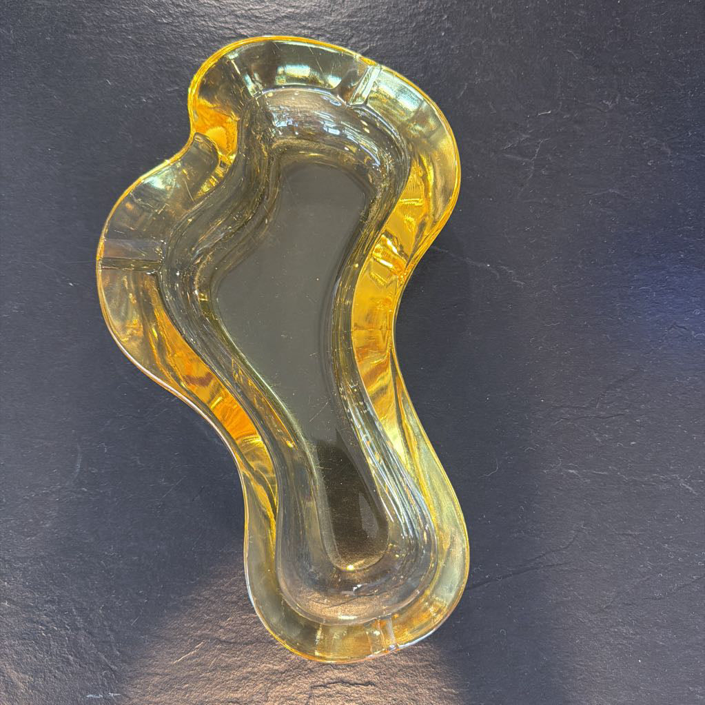 An abstract, wavy-shaped clear glass dish with a warm amber-yellow tint, photographed on a dark textured surface.