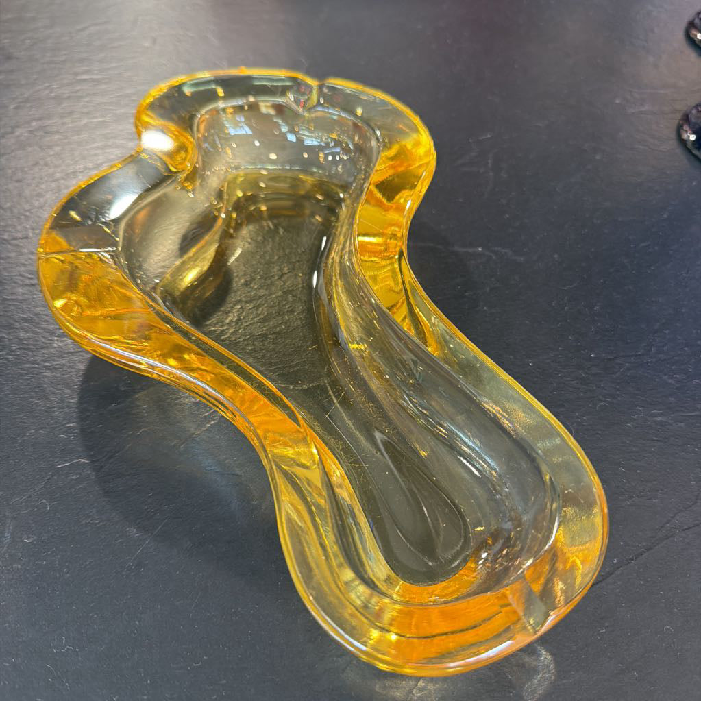 A translucent amber yellow glass ashtray with a wavy, organic shape, photographed on a dark gray textured surface.