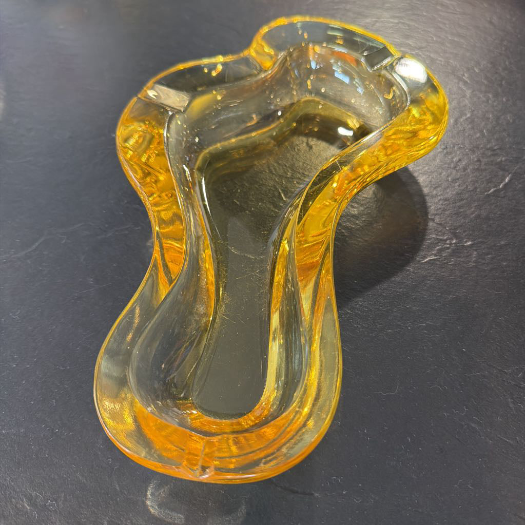A translucent amber-yellow glass dish with an organic, wavy shape, photographed on a dark gray textured surface.