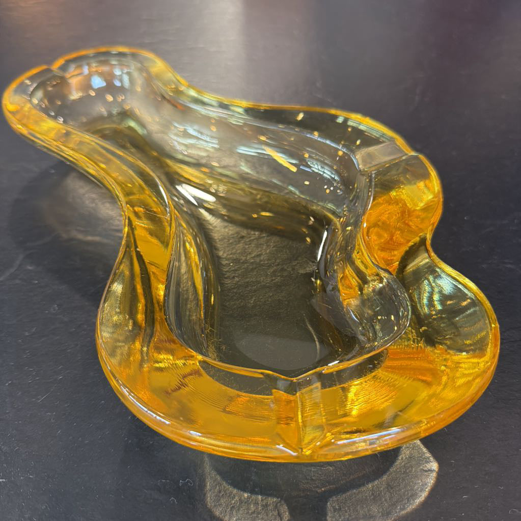 A translucent amber-yellow glass ashtray with an organic, wavy shape, photographed on a dark reflective surface.