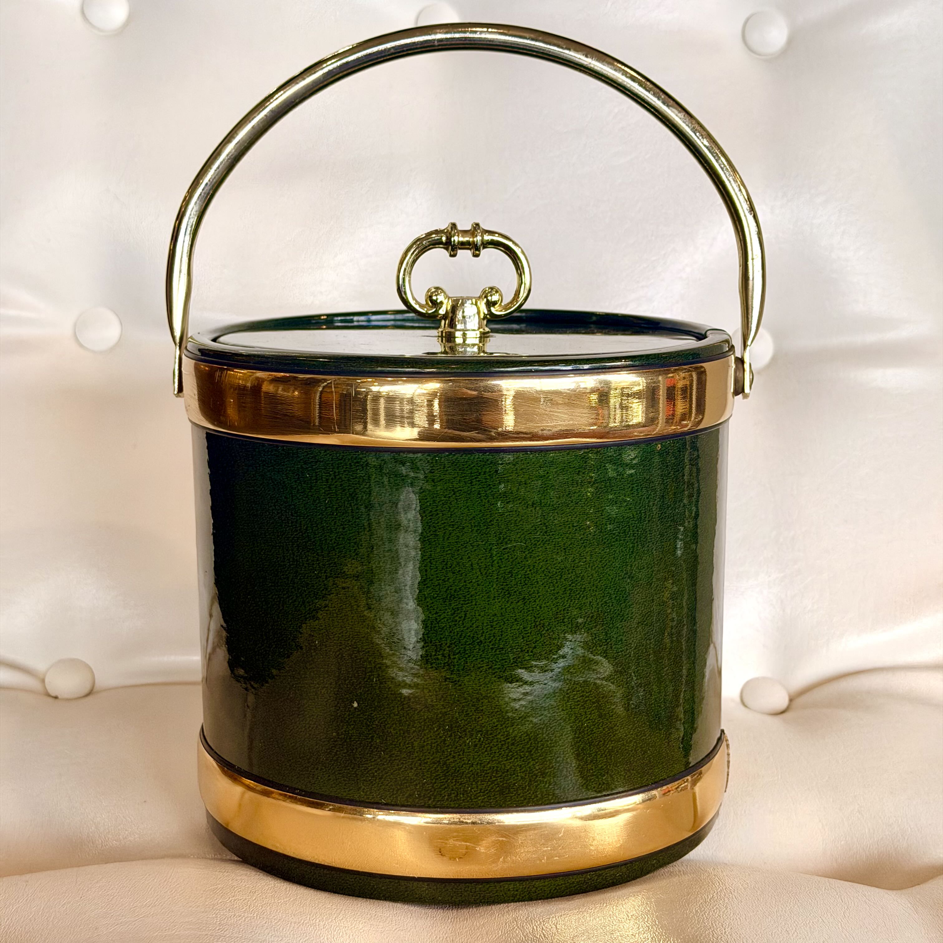 A vintage-style cylindrical bucket bag in glossy dark green with polished gold-tone metal trim, handle, and ring detail, photographed against a cream tufted fabric background.