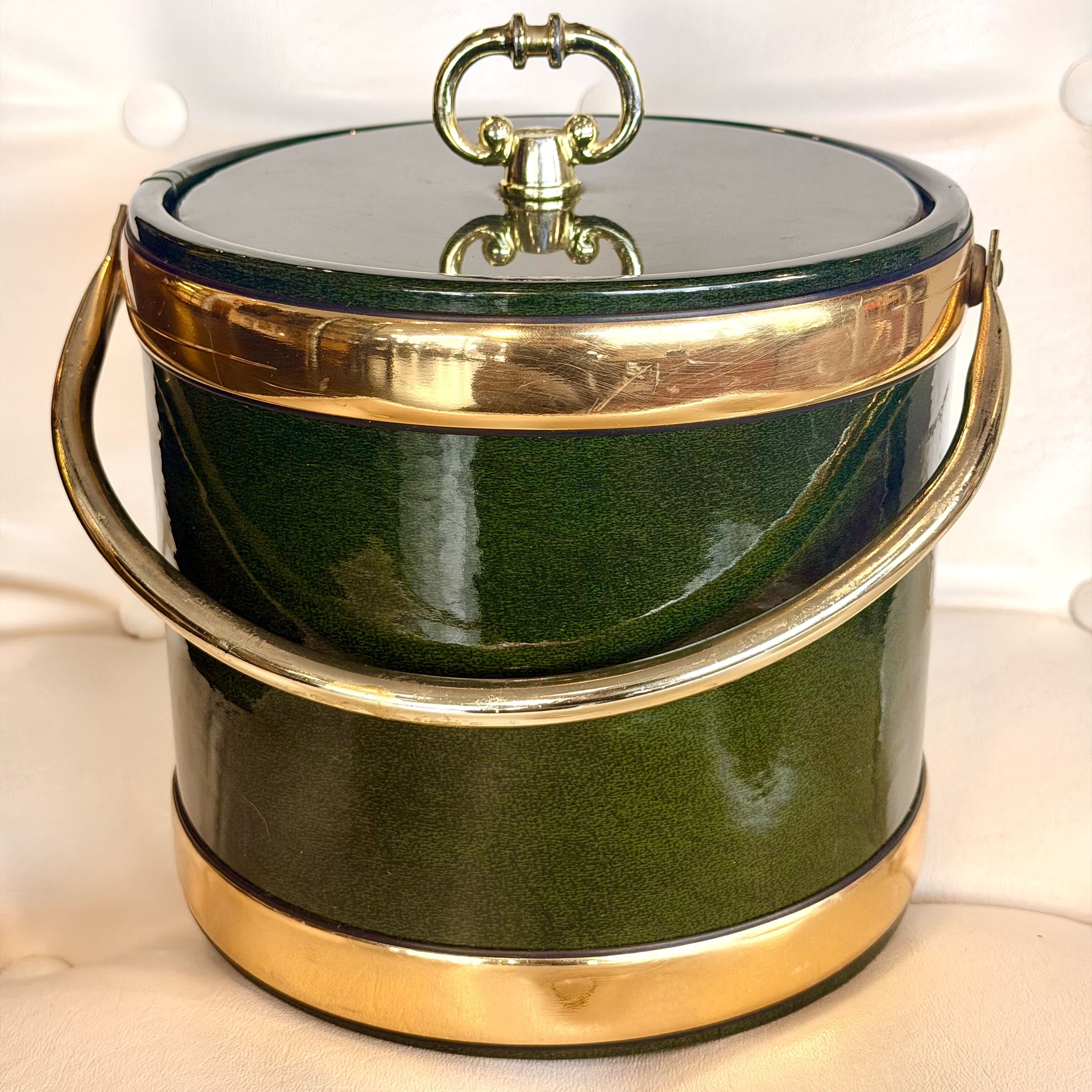 A vintage green enamel ice bucket with polished gold-tone metal trim, a lid handle, and a side carry handle, set against a light cream fabric background.