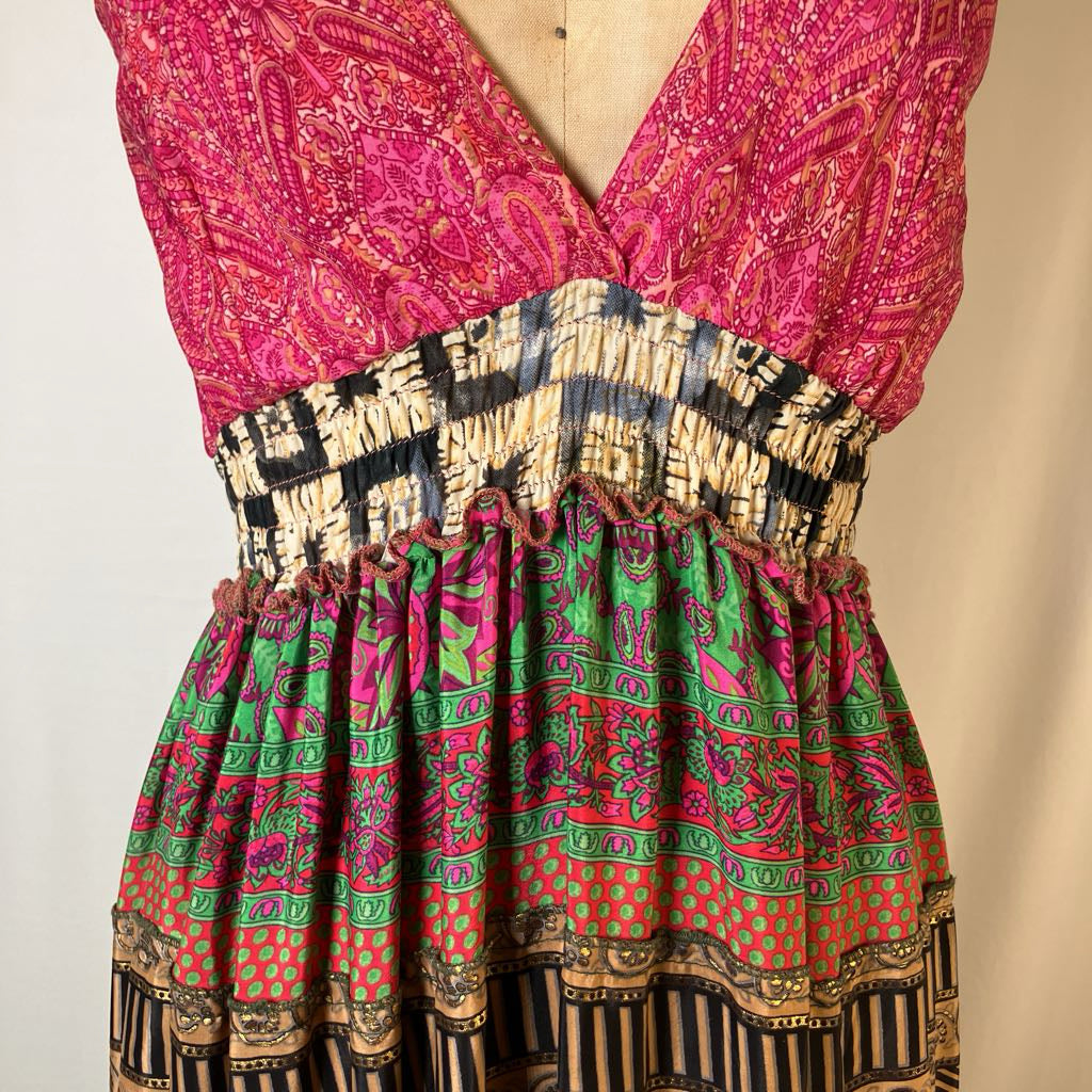 A sleeveless V-neck dress displayed on a mannequin torso, featuring a bright pink paisley bodice, a black-and-beige patterned smocked waistband, and a gathered green, pink, orange, and black patterned skirt.