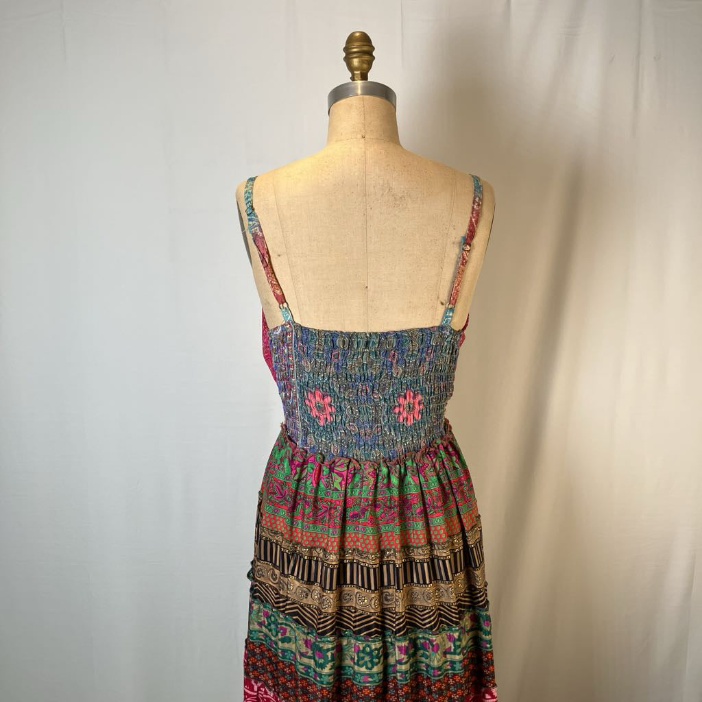 A sleeveless smocked dress displayed on a fabric mannequin torso against a light neutral backdrop. The dress has a multicolor bohemian pattern with pink, green, blue, black, and beige tones, featuring thin patterned shoulder straps.