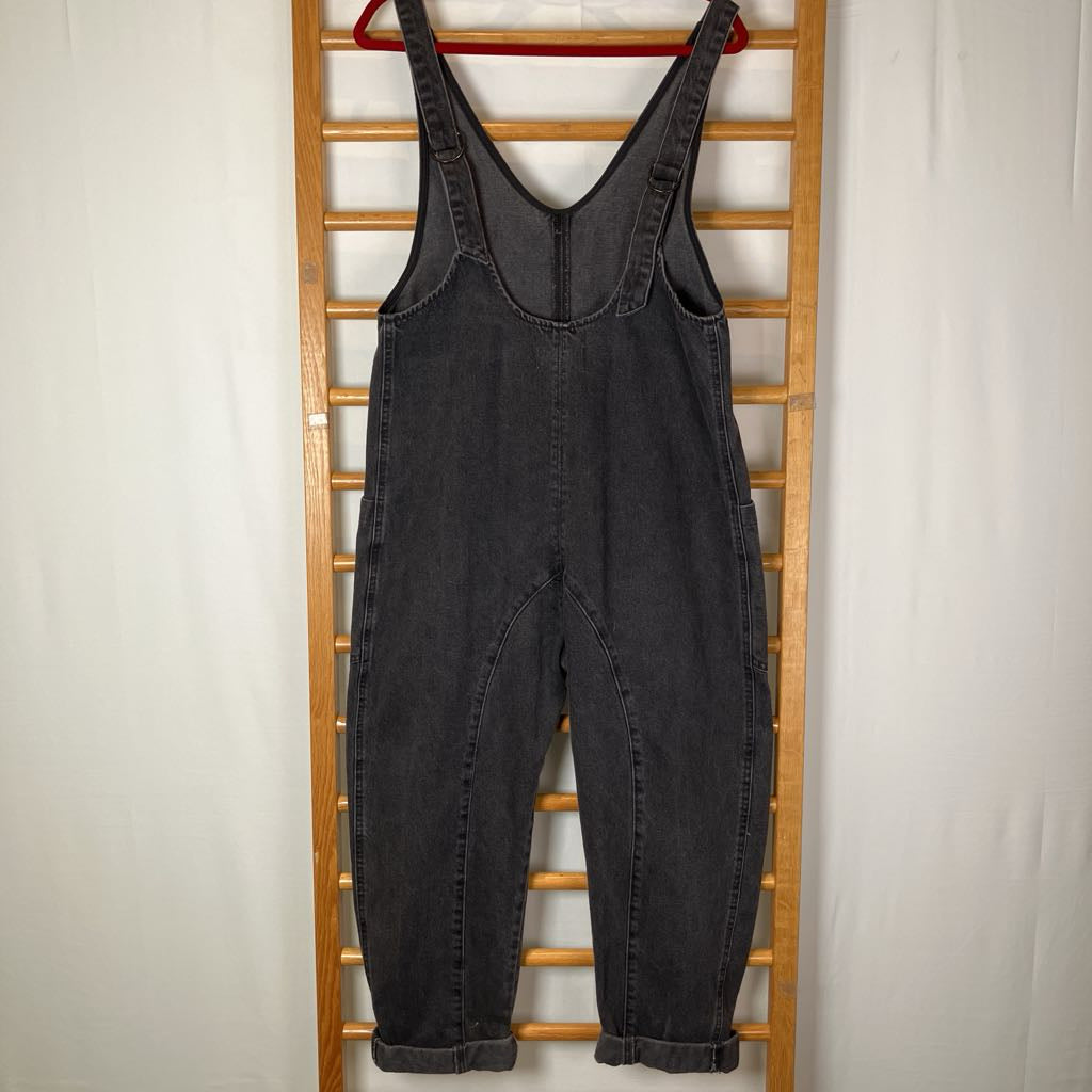 A pair of dark gray washed denim bib overalls hanging on a red hanger from a wooden ladder rack against a white fabric backdrop.