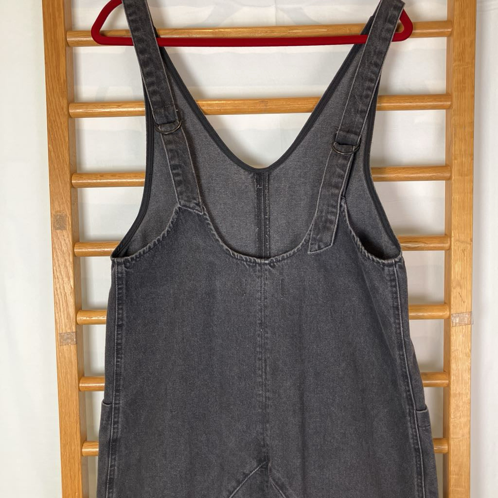 A dark gray denim overall jumpsuit hanging on a red hanger against a wooden ladder-style backdrop.