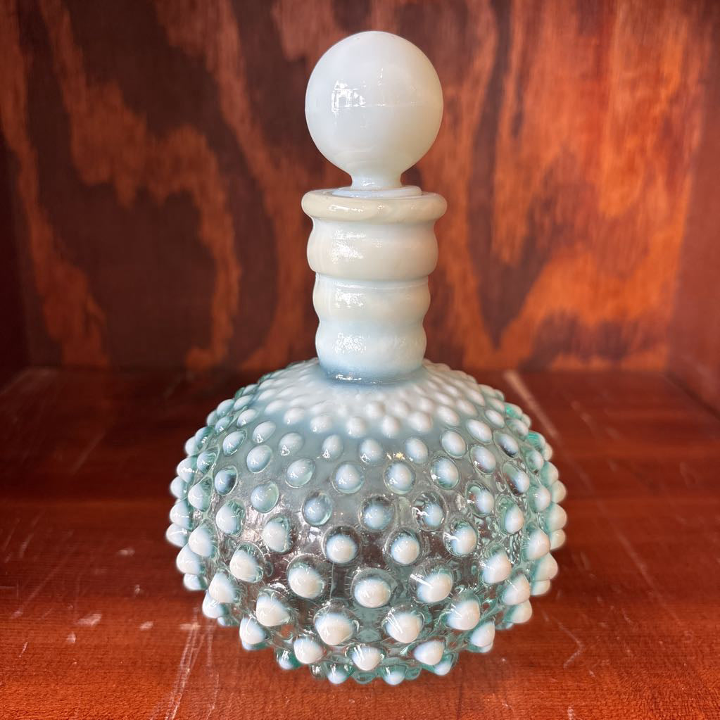 A clear and milk hobnail glass perfume bottle with a stopper in a wooden cabinet.