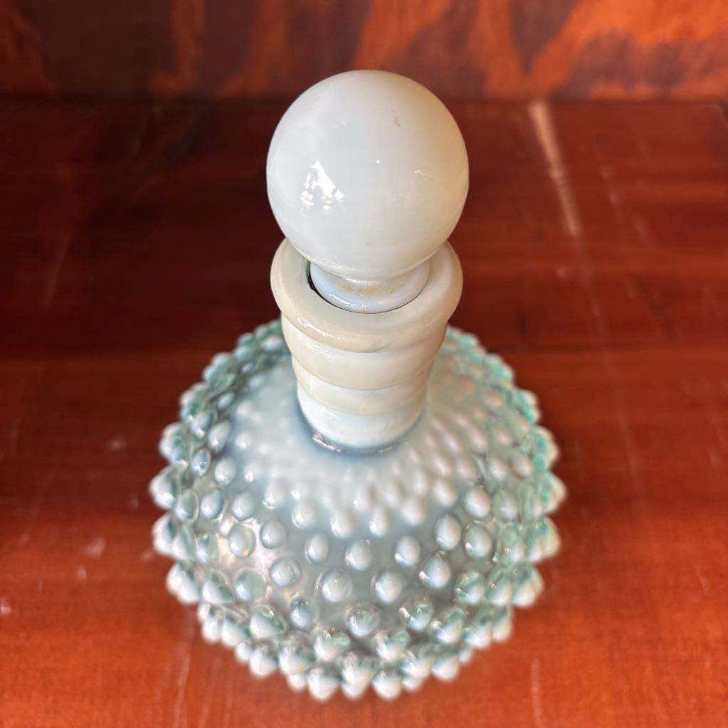 A clear and milk hobnail glass perfume bottle with a stopper in a wooden cabinet.
