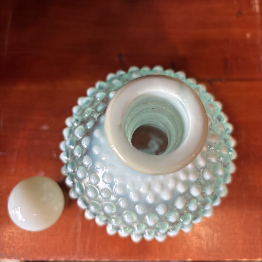A clear and milk hobnail glass perfume bottle with a stopper in a wooden cabinet.