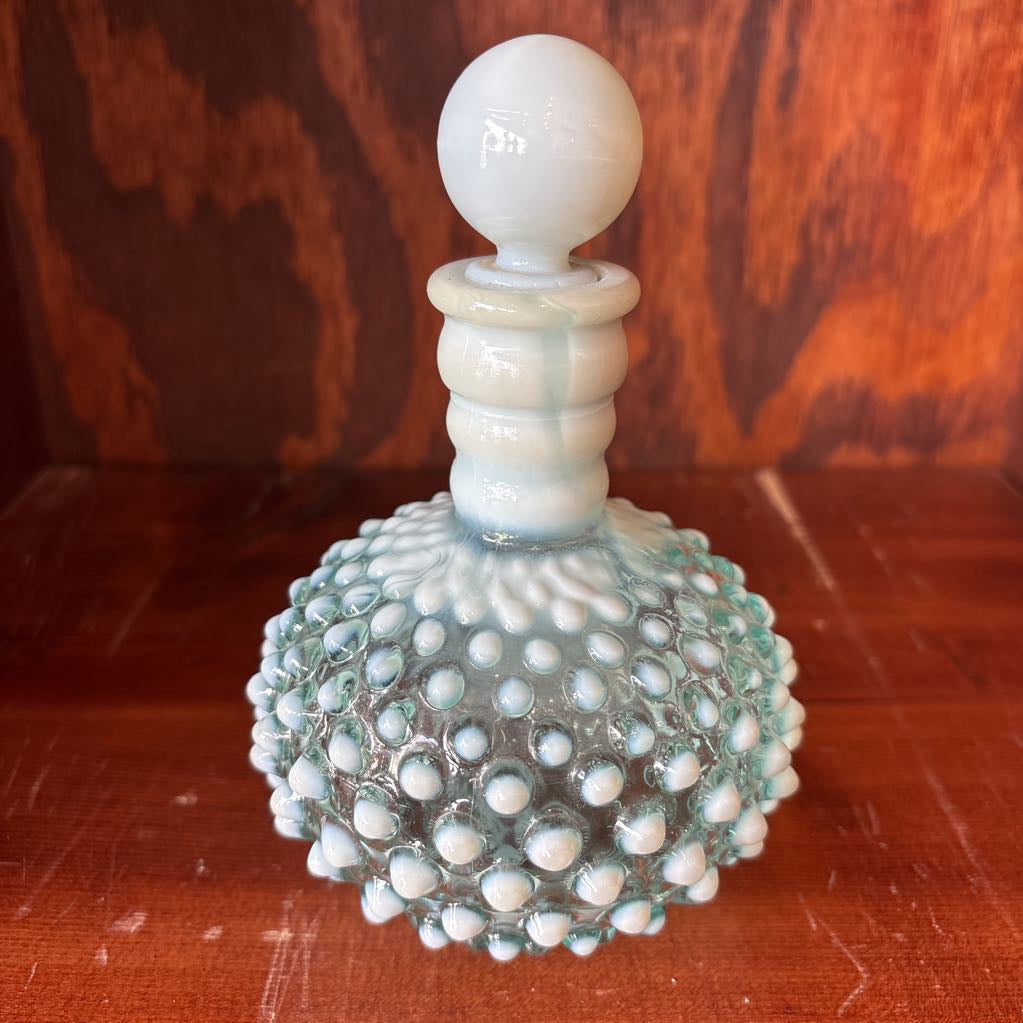 A clear and milk hobnail glass perfume bottle with a stopper in a wooden cabinet.
