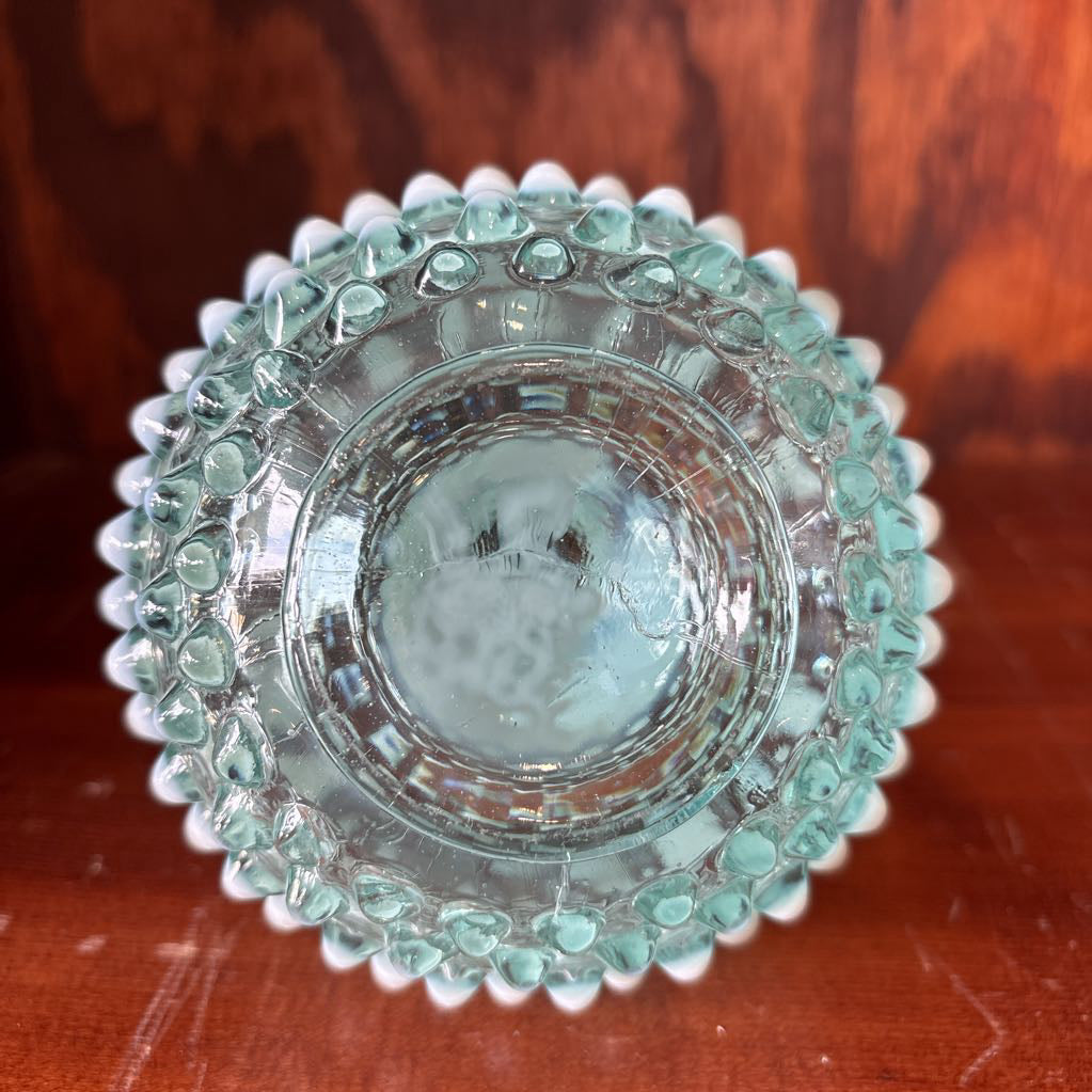 A clear and milk hobnail glass perfume bottle with a stopper in a wooden cabinet.