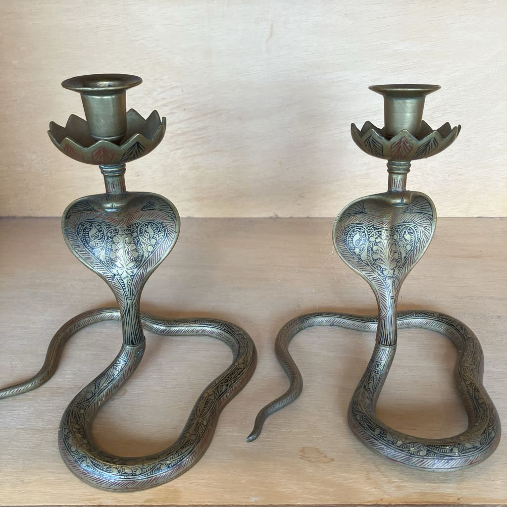 A pair of brass snake candlesticks with detailed engravings and lotus-shaped candle holders, displayed on a light wood surface against a wooden background.