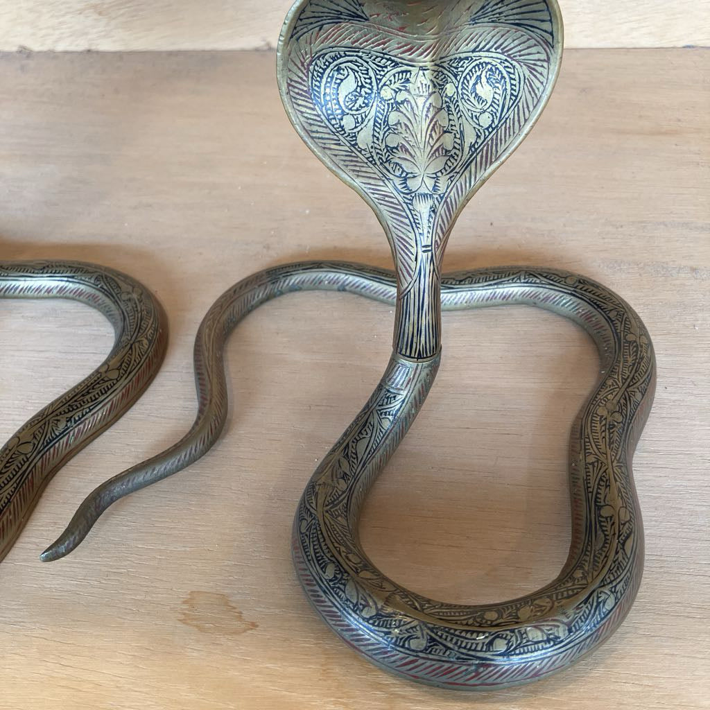 An ornate metal snake-shaped candleholder with intricate engraved patterns, displayed on a light wood surface.