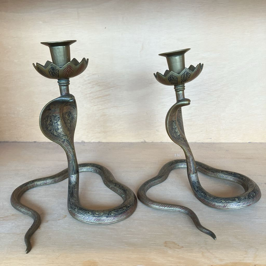 A pair of decorative metal candlestick holders shaped like coiled snakes, photographed on a light wood surface against a matching wood background.