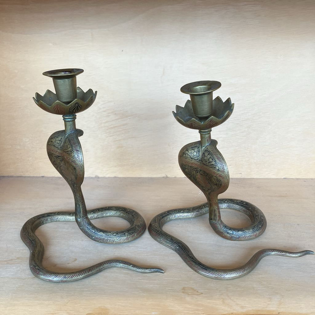 A pair of brass snake-shaped candlesticks with lotus flower cup holders, displayed on a light wood surface against a wooden background.