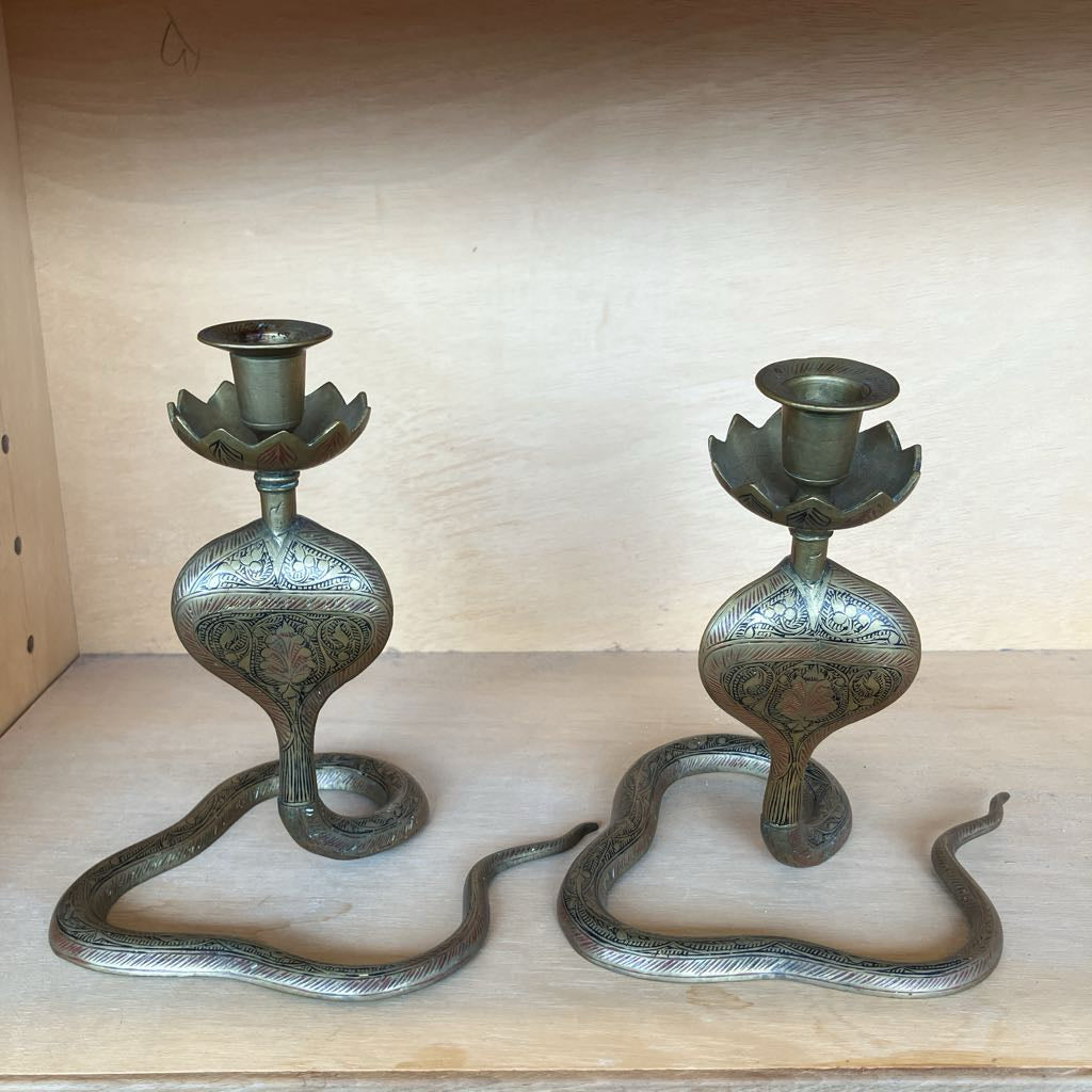 A pair of ornate snake-shaped metal candlesticks with lotus flower cup holders, displayed on a light wood shelf.
