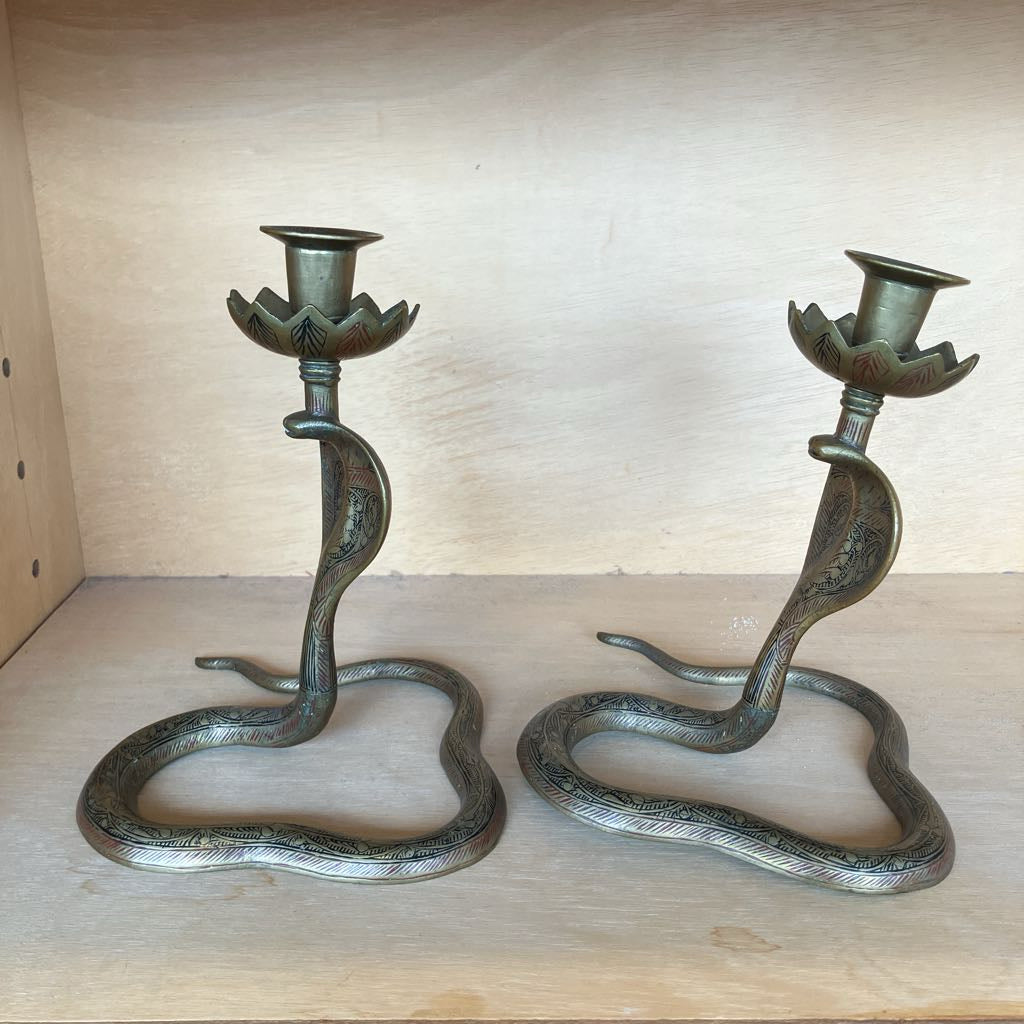 A pair of snake-shaped candlestick holders with lotus flower cup tops, shown on a light wood shelf against a wooden backing.