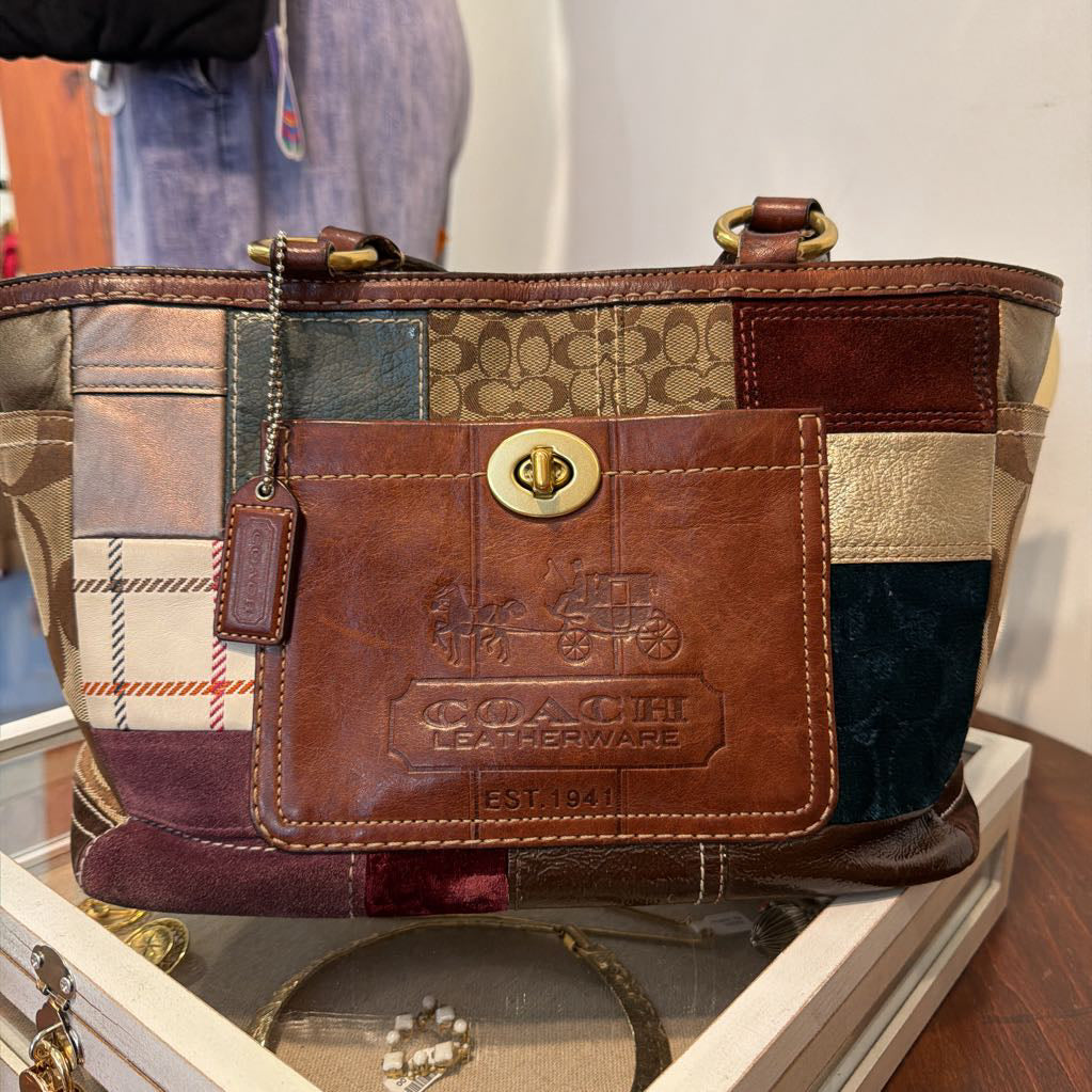 A Coach patchwork leather tote bag with a brown leather front pocket embossed with the Coach logo, displayed on a glass case.