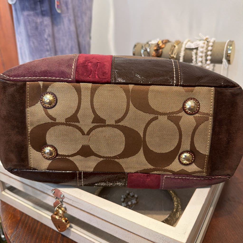 A close-up of the bottom corner of a Coach handbag, showing the classic brown and beige monogram canvas, dark brown leather trim, and gold-tone hardware studs, resting on a white jewelry box.