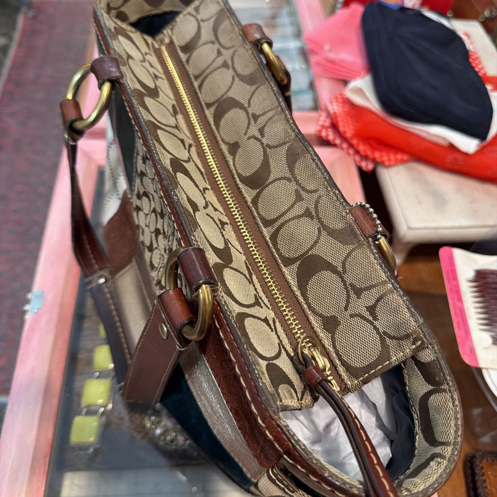 A close-up of a Coach handbag with the brand's signature monogram canvas, brown leather trim, and gold-tone hardware, displayed on a glass shelf with other items in the background.