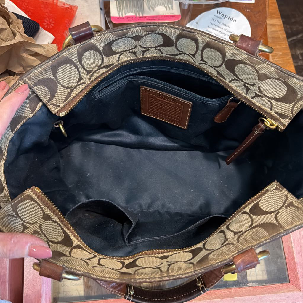 An open Coach handbag shown from the inside, featuring the brand's signature monogram canvas, brown leather trim, and a dark blue fabric lining.